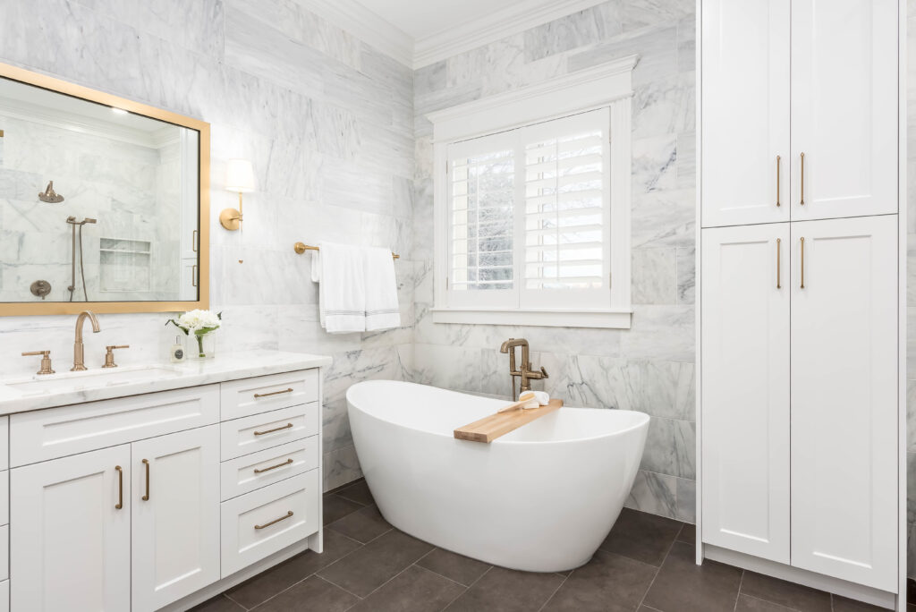 Primary bathroom remodel featuring a freestanding soaking tub, full height marble tile walls, white shaker vanity, and brushed brass fixtures.