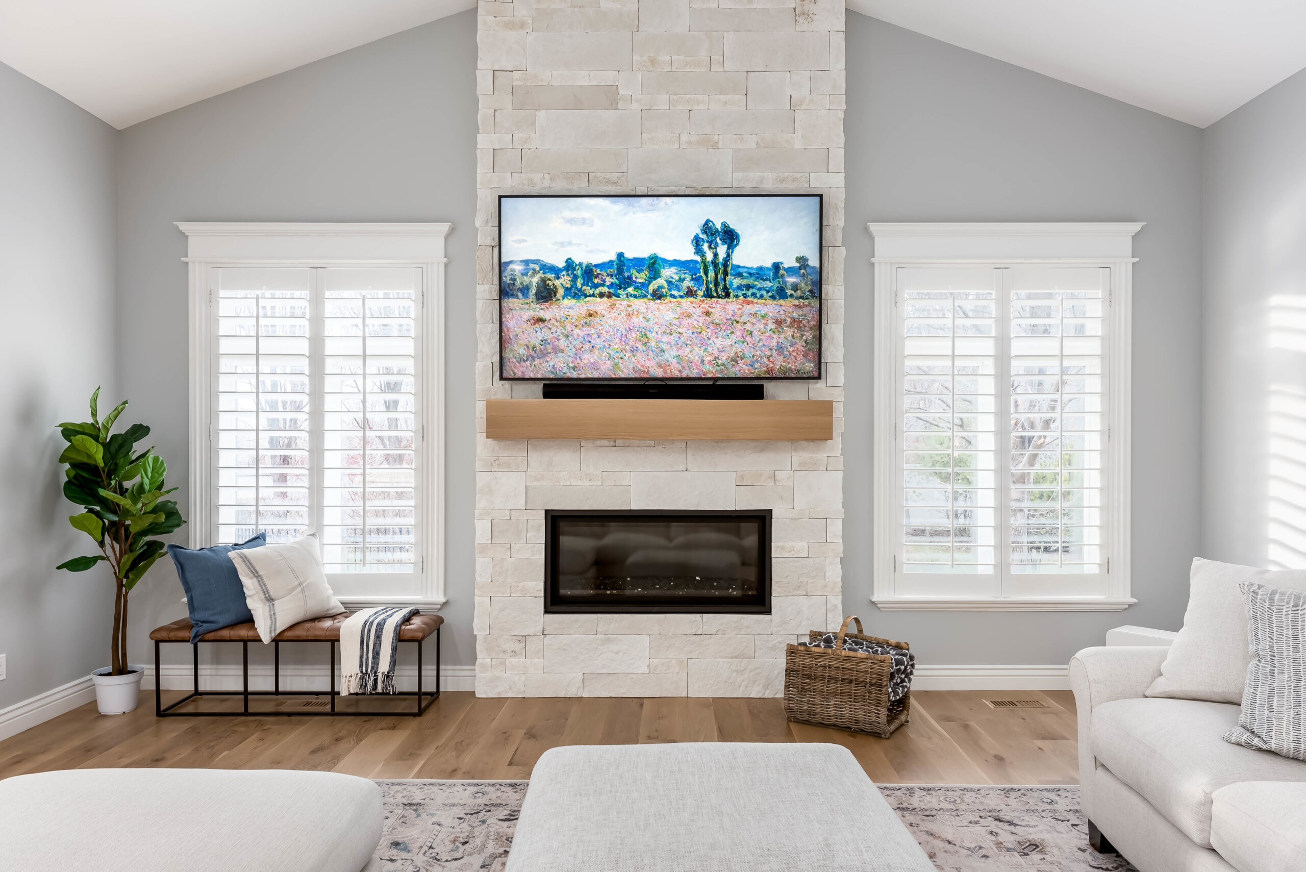 Bright modern living room featuring a floor to ceiling stone fireplace, mounted flat screen TV, white plantation shutters, and light hardwood flooring.