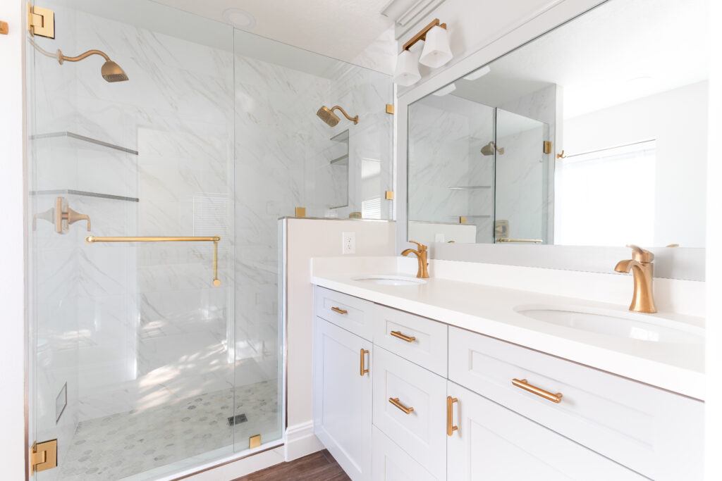 Primary bathroom remodel featuring double sink vanity, white shaker cabinetry, brushed brass hardware, and frameless glass marble shower.