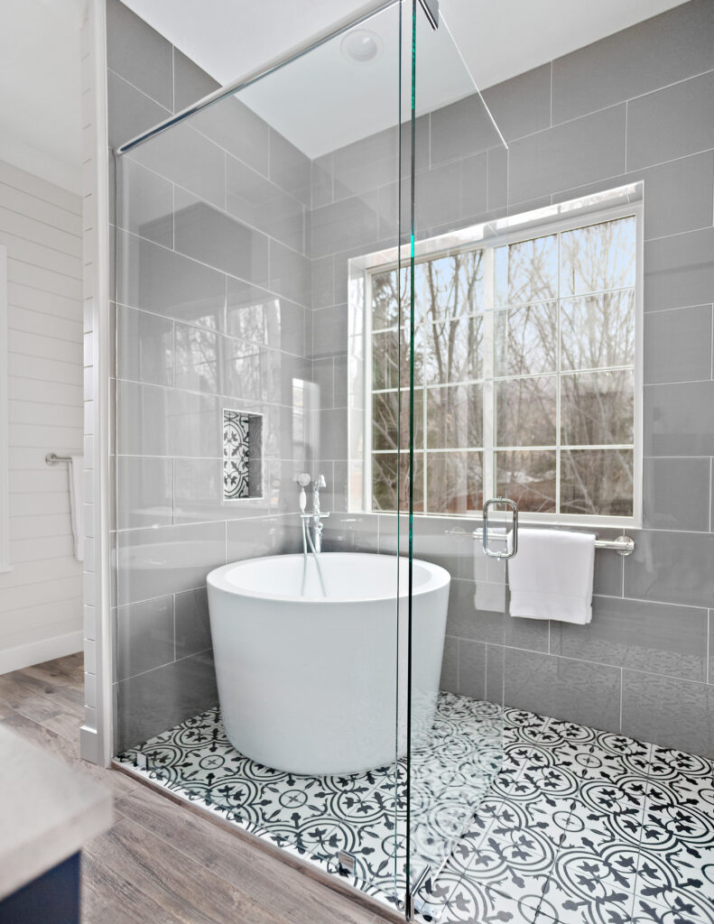 Frameless glass walk in shower with gray subway tile walls, chrome rain shower head, handheld sprayer, and patterned tile shower floor.
